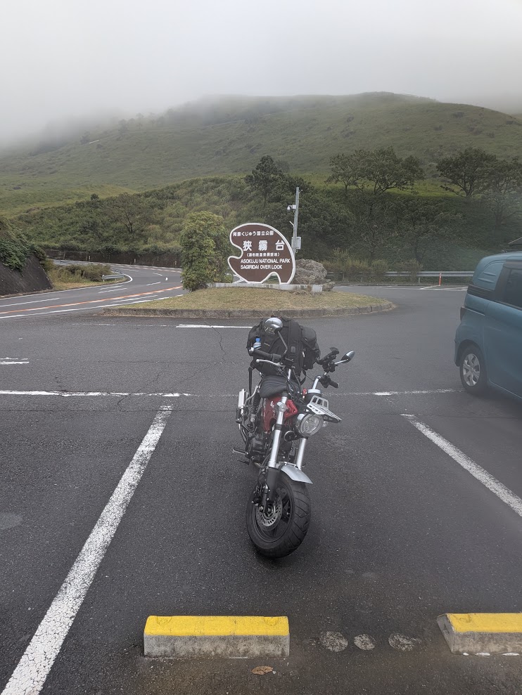 狭霧台の看板とDAX125。山に霧がかかっていて、路面が濡れている