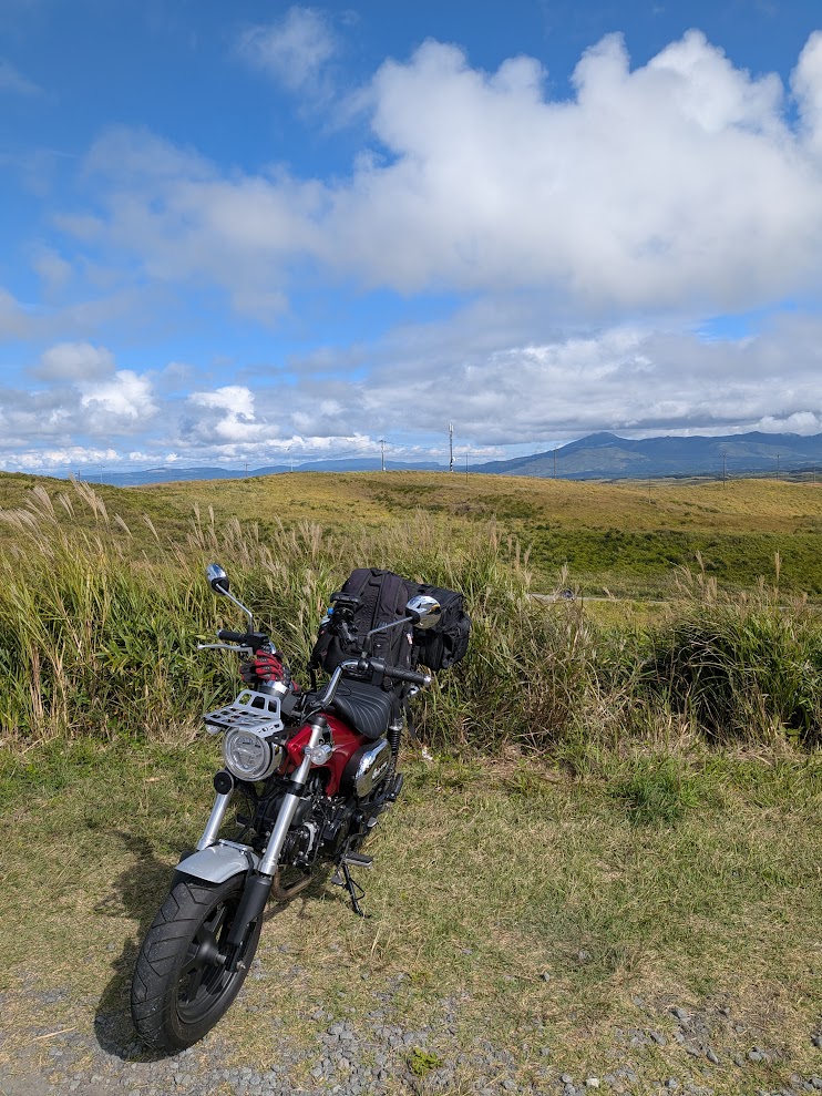 阿蘇の草原。ススキと青空、遠くに山並み。路肩にDAX125