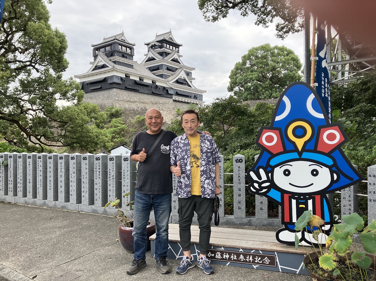 加藤神社の看板の前で、本人と上野さんが並んで写っている。背景に熊本城の天守閣