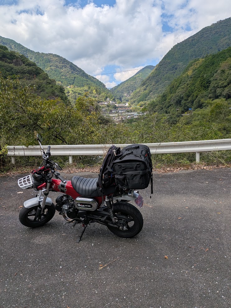 四国山地の谷。ガードレールの向こうに山あいの集落が見える。路肩にDAX125